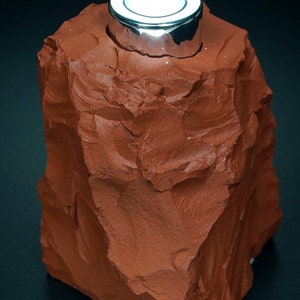 May include: A small, rough, red-brown rock with a silver metal top. The rock is shaped like a mountain and the metal top is a cylinder with a smooth, shiny surface.