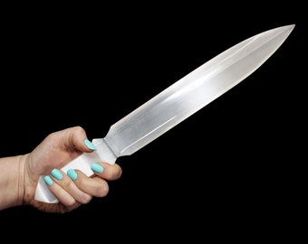 Puede incluir: Una espada de cristal de selenita transparente con un acabado liso y pulido. La espada está sostenida en una mano con esmalte de uñas azul claro.