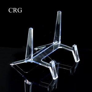 May include: Clear acrylic display stand with a three-legged design. The stand is transparent and features the letters "CRG" in the upper left corner. The stand is designed to hold plates or other items.