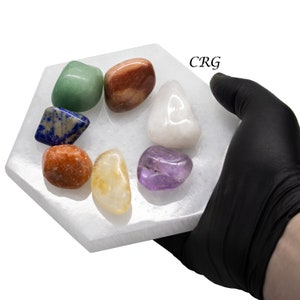May include: A white selenite charging plate with seven chakra stones: red, orange, yellow, green, blue, indigo, and violet. The stones are arranged in a circle on the plate. The plate is being held by a gloved hand. CRG is written on the plate.