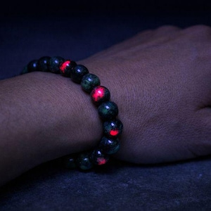 May include: A dark bracelet made of round, dark green beads with red glowing accents. The bracelet is worn on a person's wrist, with the hand and arm visible against a dark background.