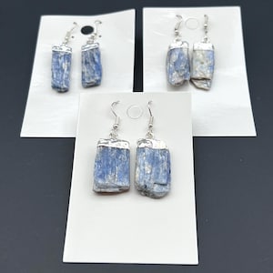 May include: Three pairs of earrings with blue kyanite stones. Each stone is rectangular and has a silver-colored metal cap and a silver hook. The earrings are displayed on white cards against a dark background.