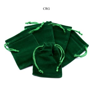 Puede incluir: Un grupo de siete bolsitas de terciopelo verde con cordón, apiladas y dispuestas sobre un fondo blanco. Las bolsitas están hechas de un tejido suave y mullido y tienen una textura lisa y sedosa.