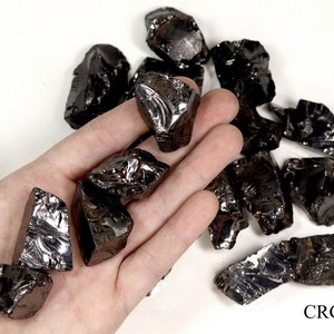 May include: A collection of black obsidian rough stones, some with a metallic sheen, on a white surface. The stones are various sizes and shapes, with some having sharp edges. The stones are arranged in a loose pile, with some overlapping. The stones are in the palm of a hand, for scale.