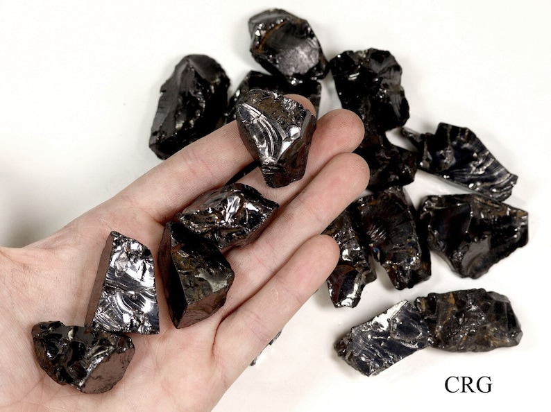 May include: A collection of black obsidian rough stones, some with a metallic sheen, on a white surface. The stones are various sizes and shapes, with some having sharp edges. The stones are arranged in a loose pile, with some overlapping. The stones are in the palm of a hand, for scale.