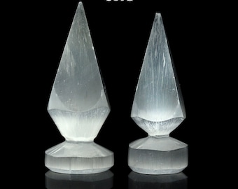 May include: Two white selenite crystal points with a pointed top and a rounded base. The crystals are on a black background.