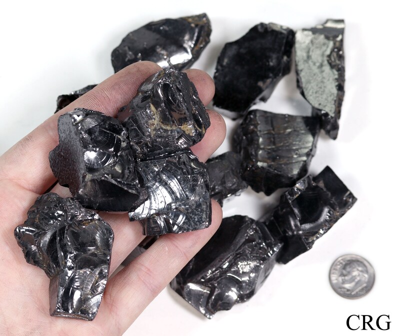 May include: A collection of black obsidian rough stones, some with a reflective surface, in a hand. A US dime is included for scale.