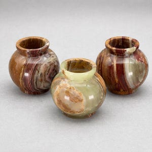 May include: Three small, decorative onyx vases. The vases are round with a wide opening and a short neck. The colors include shades of brown, red, and green, with natural veining patterns. The vases are displayed on a light gray surface.