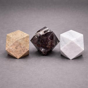 May include: Three geometric stone sculptures. The first is a light brown cube with angled facets. The second is a dark maroon diamond shape with white veining. The third is a white marble cube with angled facets.
