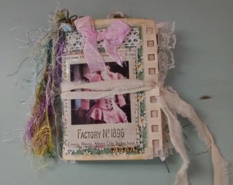 Puede incluir: Un diario hecho a mano con un lazo rosa y ribete de encaje. El diario presenta páginas con imágenes y texto vintage, incluyendo "FACTORY NO. 1896". La portada está adornada con un delicado lazo rosa y encaje, con hilos y cintas de colores.