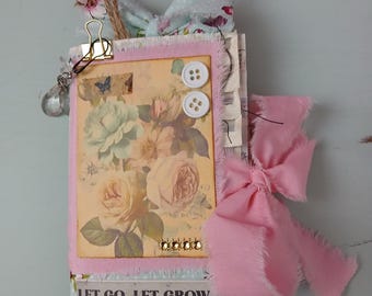Puede incluir: Un diario hecho a mano con un diseño floral en la portada, con rosas y una mariposa. El diario está adornado con una cinta rosa, botones y el texto "LET GO, LET GROW". Un clip dorado y un dije transparente añaden detalles.