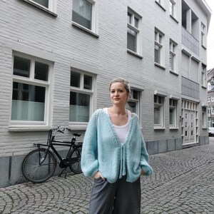 May include: A light blue, open-front cardigan with long sleeves and a tie closure. The cardigan is worn over a white top and paired with gray pants. A black bicycle is parked on a cobblestone street in front of a light gray building.
