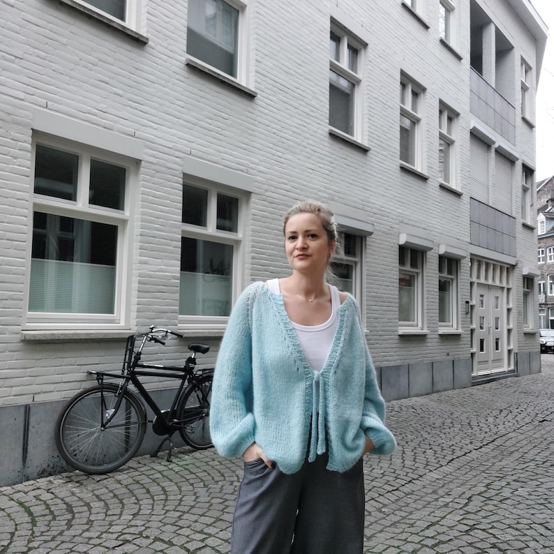 May include: A light blue, open-front cardigan with long sleeves and a tie closure. The cardigan is worn over a white top and paired with gray pants. A black bicycle is parked on a cobblestone street in front of a light gray building.