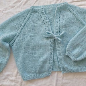 May include: A light blue knitted cardigan with long sleeves and a tie closure. The baby blue sweater has a delicate, openwork design along the neckline and sleeve seams. The cardigan is made of a soft, textured yarn.