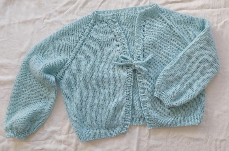 May include: A light blue knitted cardigan with long sleeves and a tie closure. The baby blue sweater has a delicate, openwork design along the neckline and sleeve seams. The cardigan is made of a soft, textured yarn.