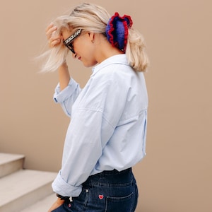 Può includere: Una donna indossa una camicia azzurra e jeans scuri con ricami di cuori rossi. I suoi capelli sono acconciati con un elastico blu e rosso. Indossa occhiali da sole con la scritta "DOLCE&GABBANA".