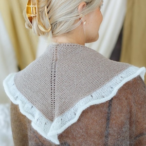 May include: A triangular knit shawl in a solid taupe color featuring a central vertical stitch detail and a delicate white ruffled lace-like border. The accessory is draped over the shoulders, showcasing a fine-gauge knit texture and soft edges.