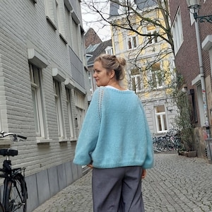 May include: A light blue knit sweater with a crew neck and long sleeves. The sweater is paired with gray wide-leg pants. The person is walking down a cobblestone street lined with buildings.