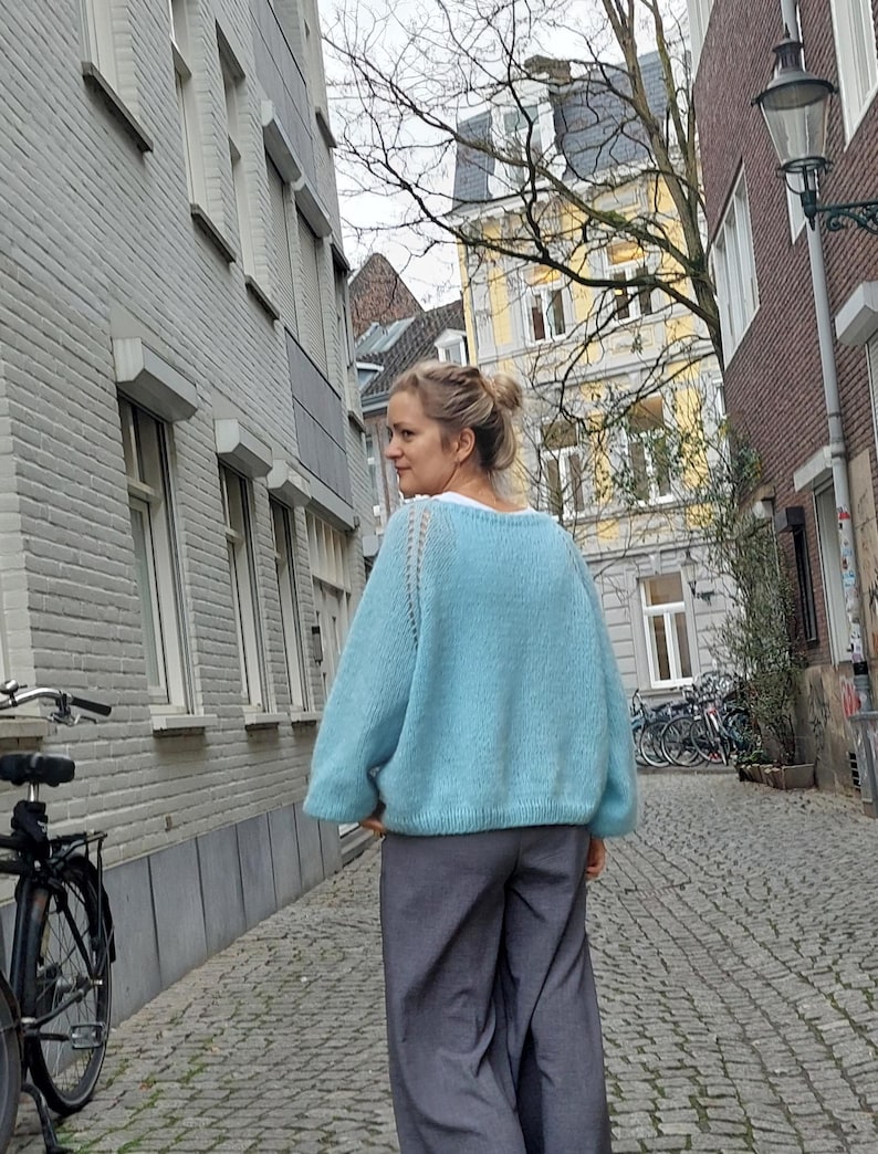 May include: A light blue knit sweater with a crew neck and long sleeves. The sweater is paired with gray wide-leg pants. The person is walking down a cobblestone street lined with buildings.