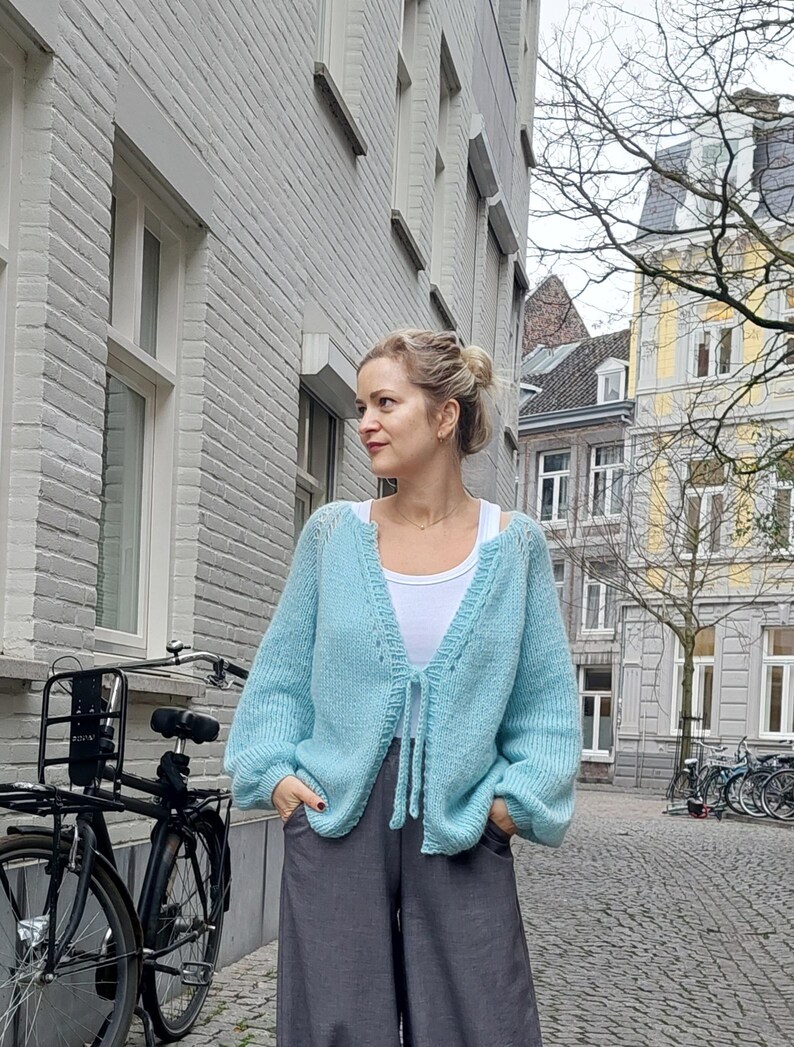 May include: A light blue, open-front cardigan with long sleeves and a tie closure. The cardigan is paired with gray pants and a white top. The image is taken outdoors, with a bicycle and buildings in the background.