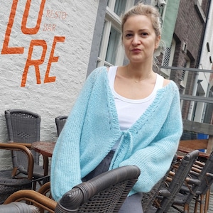 May include: A light blue, open-front cardigan with a tie closure. The cardigan is worn over a white top. The background includes a brick building with the orange text "LURE RESTO BAR". Wicker chairs and tables are in the foreground.