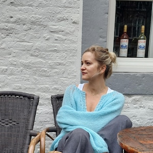 May include: A woman wearing a light blue knit cardigan and gray pants sits in a wicker chair. The cardigan has a V-neck and long sleeves. The background includes a white brick wall, a window with two bottles, and a wooden table.
