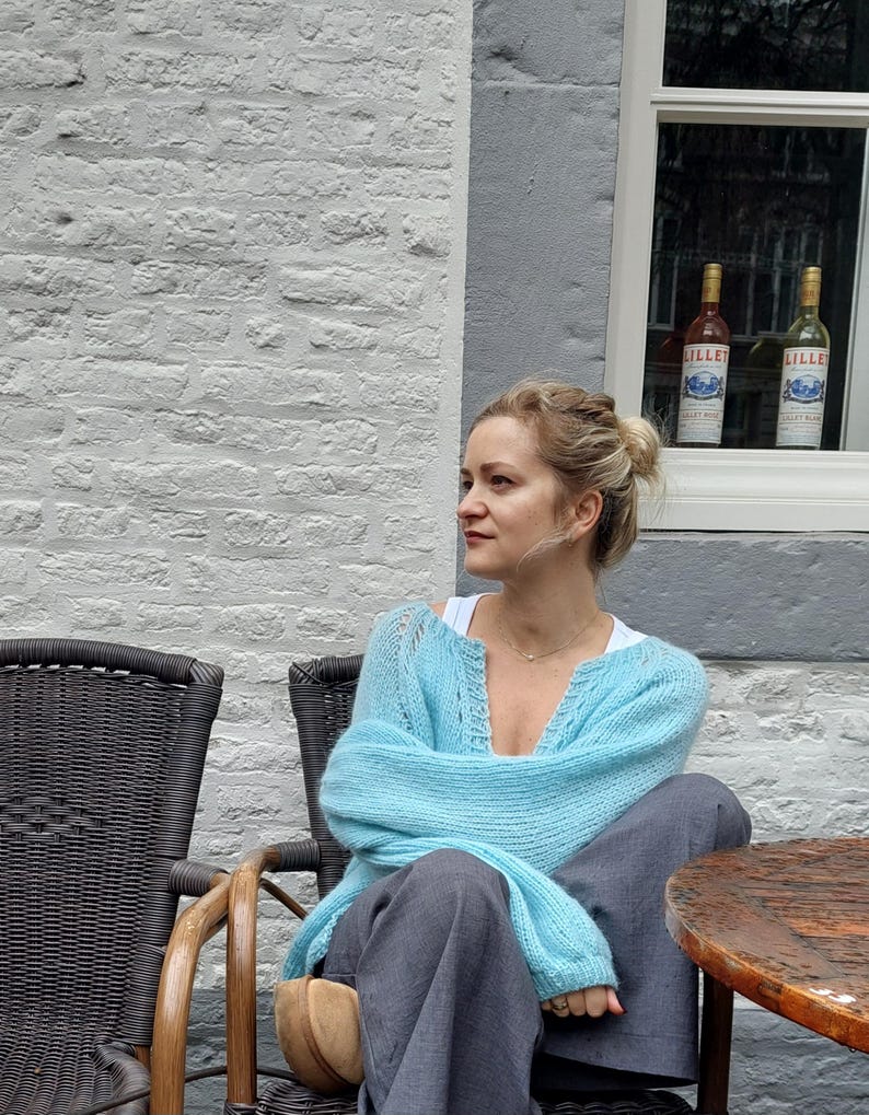 May include: A woman wearing a light blue knit cardigan and gray pants sits in a wicker chair. The cardigan has a V-neck and long sleeves. The background includes a white brick wall, a window with two bottles, and a wooden table.