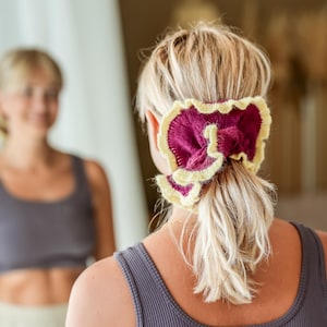 May include: A woman with blonde hair wearing a purple and yellow scrunchie in a low ponytail. The scrunchie is a large, decorative hair accessory. The woman is wearing a gray tank top. A mirror reflects the woman's face.