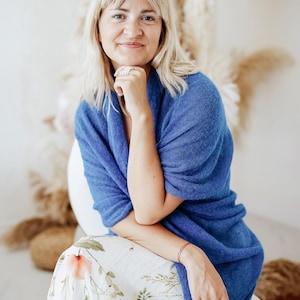 May include: A woman wearing a blue knitted shawl over a white floral skirt. She is sitting on a white chair with a gold base.