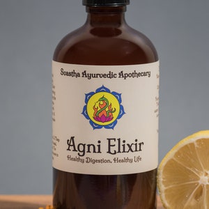 Puede incluir: Una botella de vidrio marrón con una tapa negra y una etiqueta blanca que dice "Swastha Ayurvedic Apothecary Agni Elixir Healthy Digestion, Healthy Life". La botella está sobre una superficie de madera con una rodaja de limón y clavos.