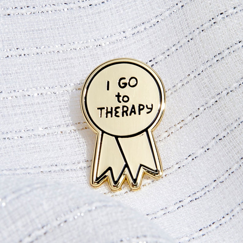 Puede incluir: Pin de esmalte de color dorado en forma de cinta con el texto "I GO TO THERAPY" en negro.