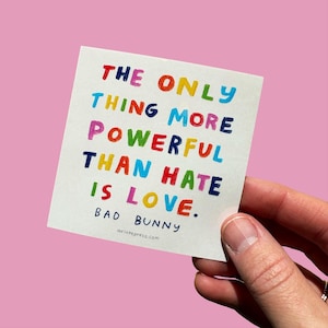 May include: A square white card with the text "THE ONLY THING MORE POWERFUL THAN HATE IS LOVE." in colorful block letters. The card also includes the name "BAD BUNNY" and the website aviatepress.com.