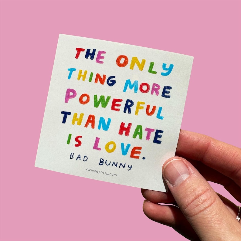 May include: A square white card with the text "THE ONLY THING MORE POWERFUL THAN HATE IS LOVE." in colorful block letters. The card also includes the name "BAD BUNNY" and the website aviatepress.com.