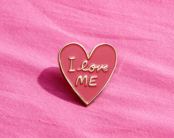 May include: A pink heart-shaped enamel pin with gold trim. The pin says "I love ME" in white script.
