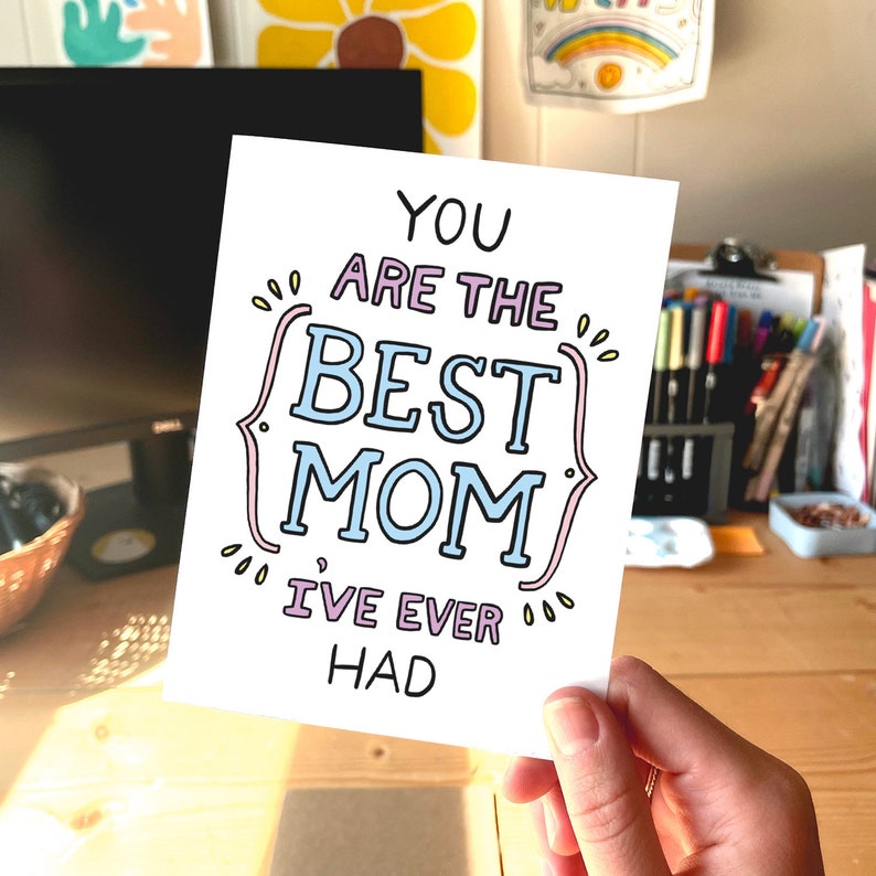 May include: A white greeting card with the text "YOU ARE THE BEST MOM I'VE EVER HAD" in a playful font. The words "BEST MOM" are in a light blue color, and the card is held in a person's hand.
