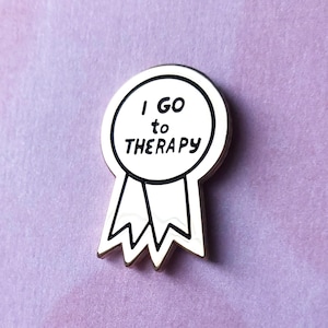 Puede incluir: Un pin de esmalte blanco con un contorno negro en forma de cinta con el texto "I GO TO THERAPY" dentro del c&iacute;rculo.
