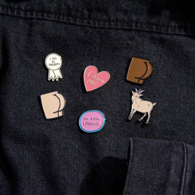 Puede incluir: Cinco pines de esmalte sobre un fondo de mezclilla negro. Los pines son: un coraz&oacute;n rosa con "Love Me" escrito en el interior, un trasero marr&oacute;n, una cabra blanca, un c&iacute;rculo rosa con "I'm Cool I Promise" escrito en el interior y una cinta dorada con "I Go To Therapy" escrito en el interior.