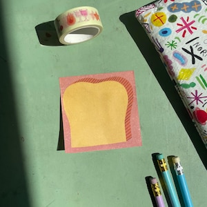 May include: A yellow sticky note with a pink border shaped like a slice of bread. The note is on a green surface with a roll of washi tape and three pencils.