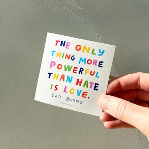 May include: A white square sticker with the text "THE ONLY THING MORE POWERFUL THAN HATE IS LOVE." in colorful block letters. The sticker also includes the name "BAD BUNNY" and the website "ariannapress.com".