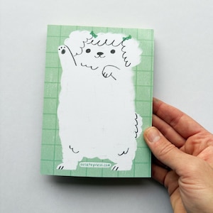 May include: A notepad featuring a cartoon illustration of a white dog with green bows, waving its paw. The dog is on a light green grid background. The notepad is held by a hand.