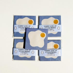 May include: A set of "Sunny Side Up" sticky notes, each with a white egg shape and a yellow yolk on a blue backdrop. The packaging displays the text "Sunny Side Up Sticky Notes - 50 Pages - AVIATEPRESS.COM".