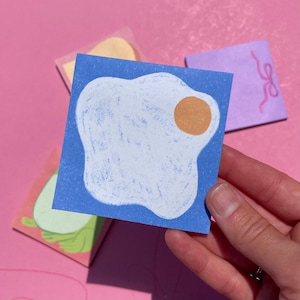 May include: A hand holding a blue square sticky note with a white fried egg illustration and a yellow yolk. Other sticky notes with illustrations are in the background. The background is pink.