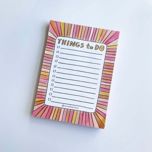 May include: A pink and orange striped notepad with the text "THINGS to DO" in gold lettering. The notepad has a smiley face at the end of the text and a blank space for writing a to-do list. The notepad has the text "@aviatepress" at the bottom.