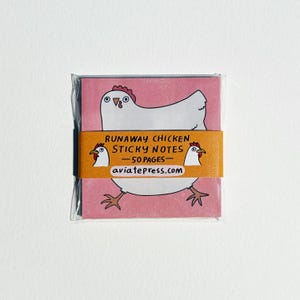 May include: A pink sticky note pad with a cartoon chicken illustration. The pad is wrapped in clear plastic and has a brown band with the text "Runaway Chicken Sticky Notes - 50 Pages - aviatepress.com".