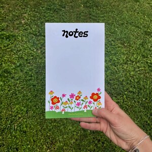 May include: A white notepad with the word "notes" in black cursive lettering. The bottom of the notepad features a green border with a row of colorful flowers and red mushrooms.