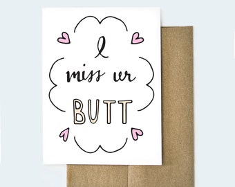 May include: A white card with a black outline of a cloud with the text "I miss ur BUTT" in black and gold lettering. There are four pink hearts around the cloud.