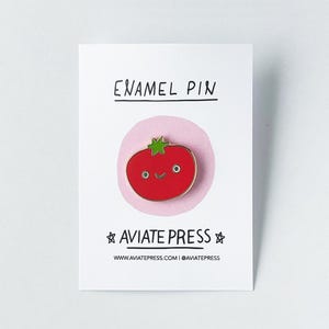 May include: A red enamel pin shaped like a tomato with a green stem and a smiling face. The pin is on a white card with the text "ENAMEL PIN" and "AVIATE PRESS" printed on it.