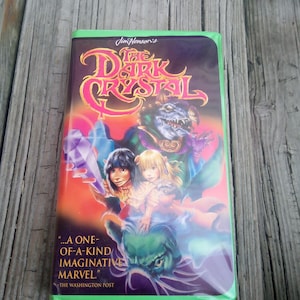 May include: A VHS tape of "The Dark Crystal" movie. The cover features a colorful illustration with the title in a stylized font. The artwork includes fantasy characters and the quote "...A ONE-OF-A-KIND IMAGINATIVE MARVEL." from The Washington Post.