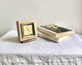 May include: A white square clock with black Roman numeral markings and gold hands. The clock is sitting on a white linen tablecloth with a stack of books behind it.