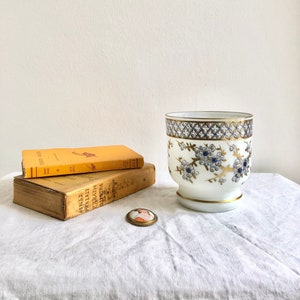 Op de afbeelding: Een witte keramische plantenpot met een blauw en gouden bloemenmotief. De pot heeft een brede, ronde basis en een licht uitlopende bovenkant. Hij staat op een wit tafelkleed met twee boeken en een klein broche.